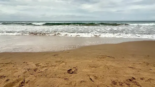 Yumuşak, altın kum, yumuşak dalgalar ve bulutlu gri gökyüzü içeren sakin ve sakin bir sahil manzarası.