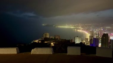 Karamsar gökyüzünün altında parıldayan kıyı şeridi boyunca şehrin ışıklarını gösteren nefes kesici bir panorama.