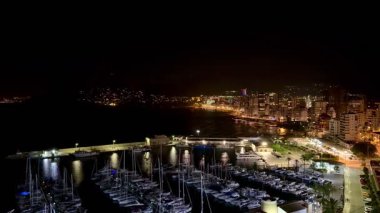 Göz kamaştırıcı bir gece limanı sahnesi aydınlık yatları ve körfez boyunca ışıl ışıl parlayan şehir silüeti.