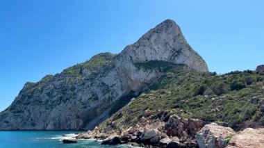 Berrak mavi gökyüzünün altında yükselen bir dağ ile eşleşen dramatik bir kayalık kıyı şeridinin nefes kesici manzarası.