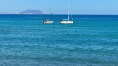 Arka planda görkemli dağlar olan sakin turkuaz sularda sallanan yelkenlilerin çarpıcı manzarası.