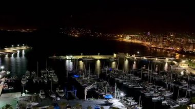 Lüks yatlarla dolu bir yat limanının göz kamaştırıcı gece manzarası ışıldayan bir şehrin siluetine vuruyor.