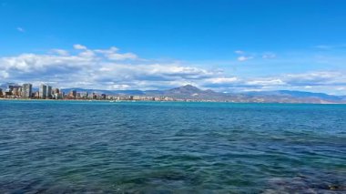 Berrak mavi gökyüzünün altında sakin suları ve görkemli dağlarıyla çarpıcı bir kıyı manzarası güzelliği yaratıyor.