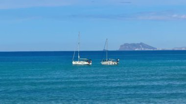 Güzel bir yelkenli sahnesi sakin sularda zarafetle sürüklenirken uzak bir ada arkaplanı manzarayı güzelleştiriyor.