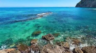 Berrak mavi gökyüzü altında canlı turkuaz suların ve kayalık oluşumların yer aldığı çarpıcı bir okyanus manzarası..