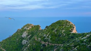 Bereketli yeşil tepelerden oluşan nefes kesici bir manzara. Pastel gökyüzünün altında sakin okyanus sularıyla güzel bir şekilde buluşuyor.