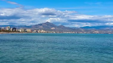 Göz kamaştırıcı plaj manzarası güzel sergileniyor. Huzurlu su, görkemli dağlar ve gökyüzünde canlı mavi bulutlar.