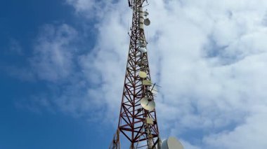 Uzun bir telekomünikasyon kulesi, antenleri açık mavi bir gökyüzüne yükseliyor, tüylü beyaz bulutlarla benekli.