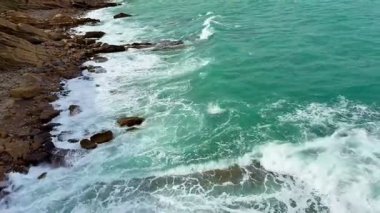 Bu hava manzarası, kayalık kıyı şeridi ve nazik dalgalarla karşılaşan çarpıcı turkuaz suları yakalar.