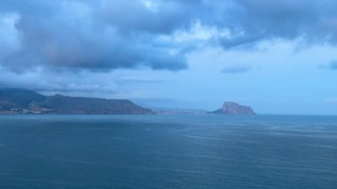 Alacakaranlıkta sakin bir deniz manzarası yumuşak dalgaların nazikçe dalgalanmasını ve yukarıdaki dramatik bulutları gösteriyor.