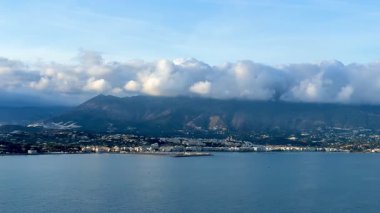 Çarpıcı manzara, yumuşak bulutlu parlak mavi gökyüzünün altında yüksek dağlar tarafından çerçevelenmiş sakin bir kıyı şeridini sergiliyor.