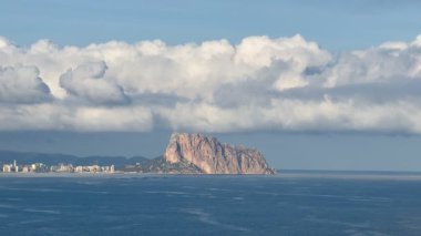 Bu kıyı manzarası bir kaya oluşumunu ve az bulutlu açık mavi gökyüzünün altında canlı bir şehir siluetini barındırır.