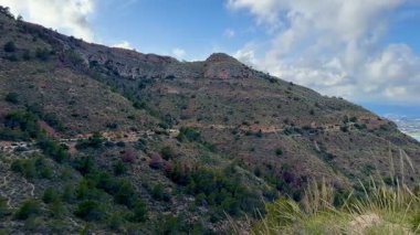 Bereketli tepelerin nefes kesici manzarası bulutlu bir gökyüzünün altındaki sakin ve dingin mavi sulara doğru iniyor.