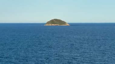 Sakin ve sakin bir ada, parlak ve berrak bir gökyüzünün altında, derin mavi denizle zarifçe çevrilidir.