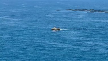 Zarif bir tekne, sakin mavi suların üzerinde zahmetsizce süzülüyor, güzel sakin kıyı manzaralarıyla çevrili.