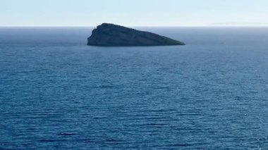 Uçsuz bucaksız okyanusun sakin manzarası, ufukta yalnız bir kaya, güzelliği ve huzuru resmediyor.