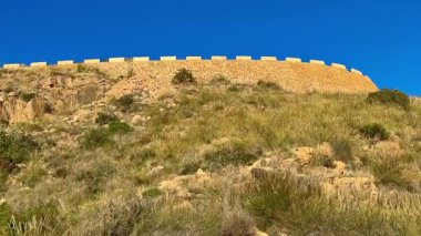 Yeşil ve açık mavi gökyüzü ile çevrili bir yamaçtaki antik taş kalenin manzarası.