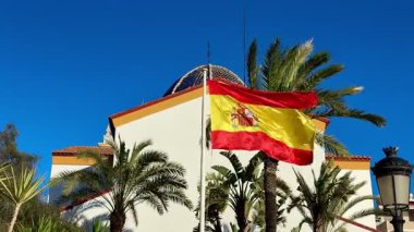 İspanyol bayrağının nefes kesici görüntüsü zarif bir şekilde palmiye ağaçları arasında sallanıyor parlak mavi bir gökyüzünün altında