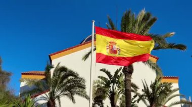 Rüzgarda dalgalanan canlı bir İspanyol bayrağı, açık mavi bir gökyüzüne ve sallanan yemyeşil palmiye ağaçlarına karşı.