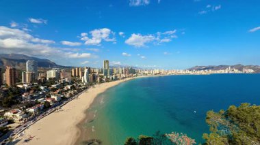 Benidorms sahilinin nefes kesici manzarası, kristal berrak sular, ve canlı bir gökyüzünün altında modern gökyüzü..