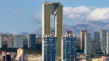 Açık mavi gökyüzünün altında şehir binaları arasında yükselen muhteşem bir gökdelenin nefes kesici manzarası.