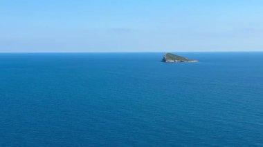 Hava manzarası güzel bir şekilde sakin bir okyanusun uzak bir adaya ve canlı bir kıyı şeridine hafifçe çarptığını gösteriyor.