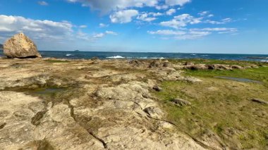 Yeşil çimenli kayalık bir sahil şeridi ve parlak, güneşli bir gökyüzünün altında derin mavi bir okyanus.