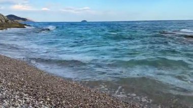 Gerçekten çarpıcı bir kıyı manzarası. Yumuşak dalgalar sergiliyor. Açık, güneşli mavi gökyüzünün altında çakıl taşlı bir plaja vuruyor.