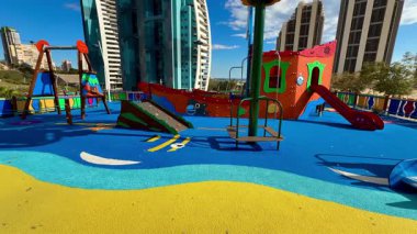 Değişik slayt ve salıncakların yer aldığı canlı ve renkli bir oyun alanı, modern bir şehrin arka planına karşı kurulmuş.