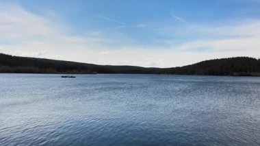 Geniş, berrak, mavi gökyüzünün altında yemyeşil ormanlarla çevrili huzurlu ve huzurlu bir göl manzarası.