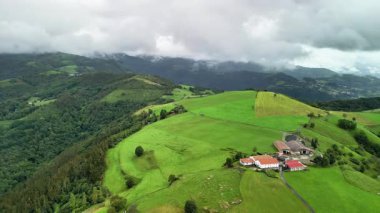 Bulutlu gökyüzünün altında büyüleyici çiftlik evlerinin yanı sıra yemyeşil tepeleri gösteren nefes kesici bir hava manzarası.
