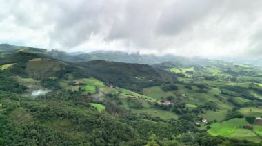 Göz kamaştırıcı bir hava manzarası yemyeşil manzaraları ve geniş bulutlu gökyüzünün altında yuvarlanan tepeleri gösteriyor.