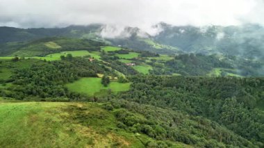 Hava manzarası canlı yeşillikler, yuvarlanan tepeler ve bulutlu bir gökyüzünün altında görkemli dağlar sergiliyor.