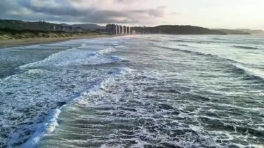 Güzel dalgalar sakin bir plaja çakılırken uzak binalar bulutlu bir gökyüzünün altında duruyor.
