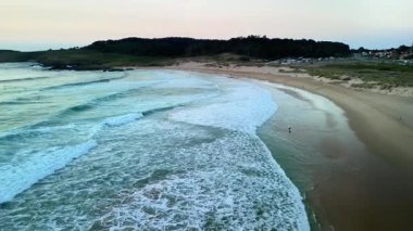 Alacakaranlıkta sakin bir sahilin havadan görünüşü yumuşak dalgaların yavaşça kıyıya vurduğu sakin bir atmosfer yaratıyor.