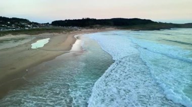 Gün batımında sakin ve huzurlu bir sahil sahnesi yumuşak dalgalar, altın kumlar ve sakin bir kıyı şeridi.