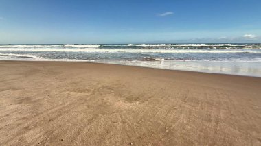 Sakin bir sahil sahnesi, yumuşak dalgalar parlak mavi bir gökyüzü ve yumuşak pamuklu bulutların altında kıyıları sarıyor.