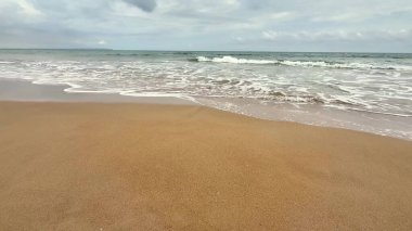 Sakin ve sakin bir sahil sahnesinde bulutlu bir gökyüzünün altında yumuşak dalgalar yavaşça kumlu kıyıya vuruyor.