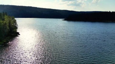 Huzurlu ve sakin bir göl, berrak mavi gökyüzünün altındaki yemyeşil tepelerin çerçevelediği güneş ışığını güzel bir şekilde yansıtır.