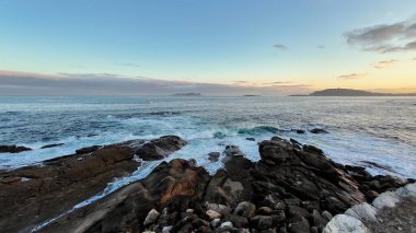 Gün batımında kayalara çarpan yumuşak dalgaların olduğu sakin bir sahil manzarası güzellikleri gözler önüne seriyor.