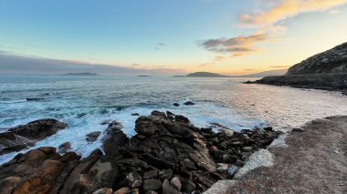 Gün batımında güzel, sakin bir kıyı manzarası, dalgaların ritmik bir şekilde kayalık bir kıyıya çarpması.
