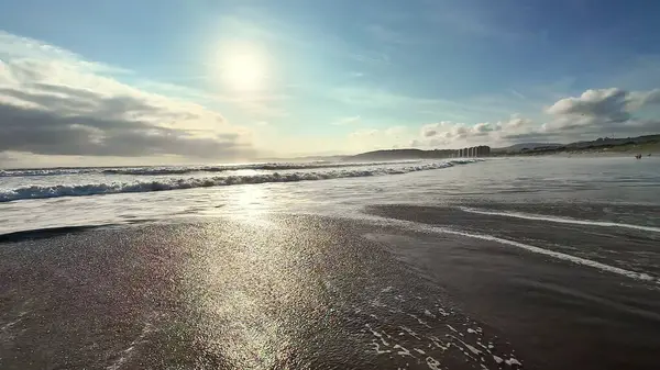 Şaşırtıcı derecede sakin bir sahil manzarası parlak güneşin altında parıldayan dalgalar sergiliyor, saf huzur ve sükuneti çağrıştırıyor.