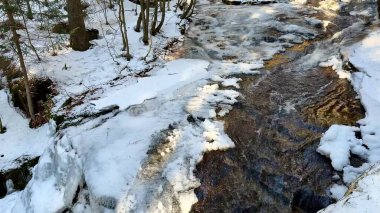 Güzel, sakin bir kış sahnesi, karla kaplı bir manzara ve ağaçlarla sarılmış, nazikçe akan bir dereyi gözler önüne seriyor.