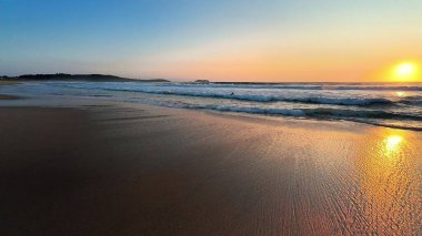 Sakin bir sahil günbatımı, sakin sularda altın yansımalar ve kıyıya vuran yumuşak dalgalar sergiliyor.