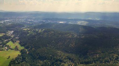 Bu hava manzarası, sisli bir gökyüzü tarafından güzel çerçevelenmiş canlı yeşil ormanları ve yuvarlanan tepeleri vurgular.