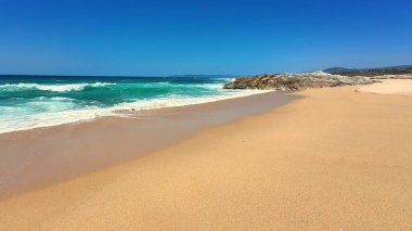 Sakin bir sahil sahnesi turkuaz dalgalar sergiliyor parlak mavi gökyüzünün altında yumuşak altın kumu yalayıp yutuyor.