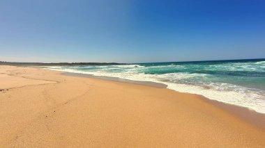 Güzel, sakin bir sahil sahnesi yumuşak altın kum, yumuşak dalgalar ve parlak mavi gökyüzü sergileniyor.