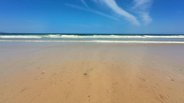 Yumuşak, sıcak kum, nazik, dingin dalgalar ve berrak mavi gökyüzünü gösteren sakin bir sahil sahnesi.