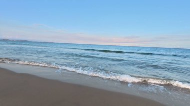 Huzurlu ve sakin bir sahil sahnesi. Yumuşak dalgaların, berrak bir gökyüzünün altında kumlu sahili usulca yalayıp yuttuğu.