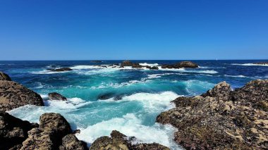 Okyanus dalgalarının nefes kesici görüntüsü berrak, mavi gökyüzünün altında kayalık çıkıntılara şiddetle çarpıyor.